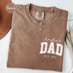 May include: Espresso-colored t-shirt with the text "Amelia's DAD EST. 2026" in white block letters. A silver necklace with a pendant rests on the shirt. The shirt is made of a soft material.