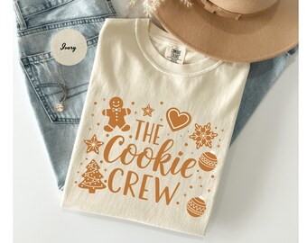 May include: Ivory-colored t-shirt with the text "THE COOKIE CREW" in a festive font, accompanied by gingerbread man, star, heart, snowflake, Christmas tree, and ornament graphics. The shirt is folded on top of blue jeans and a hat.