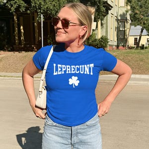 May include: A blue t-shirt with the word "LEPRECONT" in white block letters and a white shamrock graphic. The shirt is worn with light blue denim shorts and a white handbag.