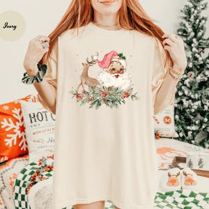 May include: Ivory-colored t-shirt featuring a festive Santa Claus and reindeer design. Santa wears a pink hat and is surrounded by greenery and berries. The shirt is part of a Christmas-themed collection.