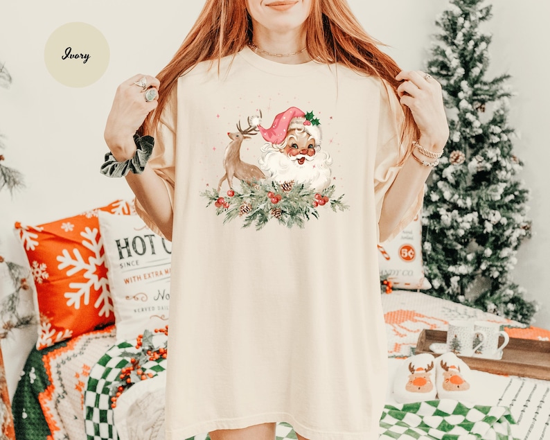 May include: Ivory-colored t-shirt featuring a festive Santa Claus and reindeer design. Santa wears a pink hat and is surrounded by greenery and berries. The shirt is part of a Christmas-themed collection.