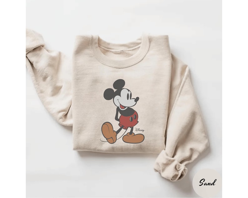 May include: Sand-colored sweatshirt featuring a classic Mickey Mouse graphic. The cartoon character is depicted in black, red, and brown, with the Disney logo. The sweatshirt has a crew neck and long sleeves.