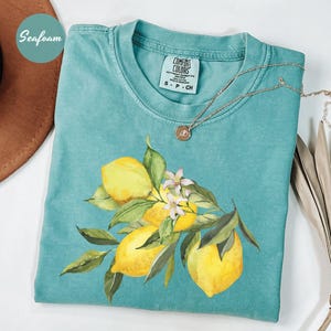 May include: Seafoam green t-shirt with a watercolor illustration of lemons and leaves. The shirt has a Comfort Colors tag and a gold necklace. A brown hat is partially visible in the upper left corner. The shirt features a lemon and floral design.