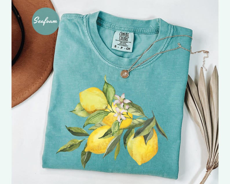 May include: Seafoam green t-shirt with a watercolor illustration of lemons and leaves. The shirt has a Comfort Colors tag and a gold necklace. A brown hat is partially visible in the upper left corner. The shirt features a lemon and floral design.