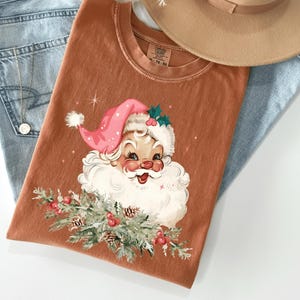 May include: A rust-colored t-shirt featuring a vintage Santa Claus graphic with a pink hat, white beard, and sprigs of greenery. The shirt is laid on top of light blue jeans and a tan hat.