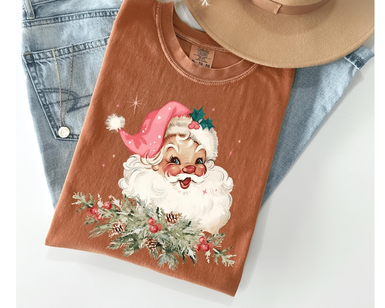 May include: A rust-colored t-shirt featuring a vintage Santa Claus graphic with a pink hat, white beard, and sprigs of greenery. The shirt is laid on top of light blue jeans and a tan hat.