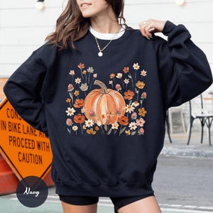 May include: A navy blue crewneck sweatshirt featuring a central graphic of a large orange pumpkin surrounded by various small wildflowers in shades of orange, yellow, and white. The design is printed on the front of the long-sleeved pullover garment.