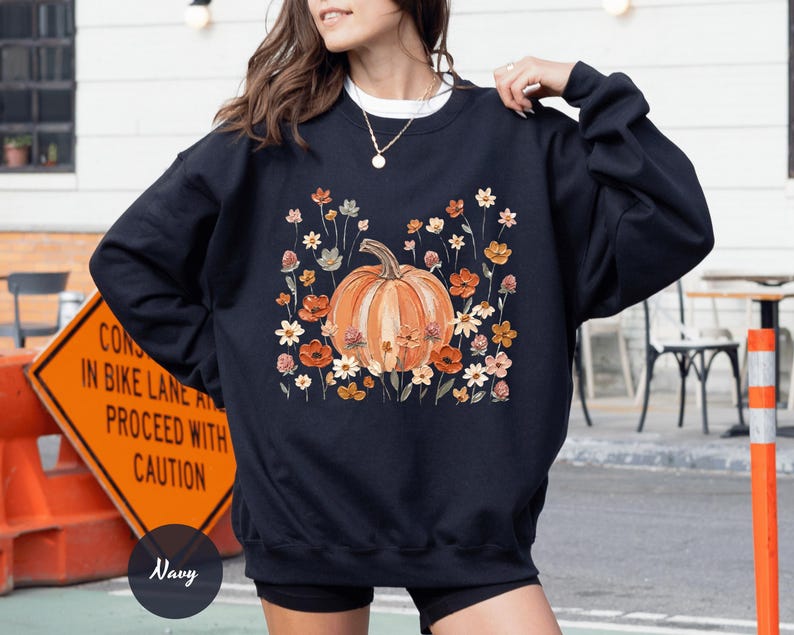 May include: A navy blue crewneck sweatshirt featuring a central graphic of a large orange pumpkin surrounded by various small wildflowers in shades of orange, yellow, and white. The design is printed on the front of the long-sleeved pullover garment.