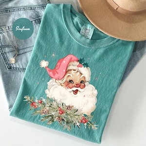 May include: Seafoam green t-shirt featuring a vintage Santa Claus graphic with a pink hat and white beard. The Santa holds a sprig of pine with red berries. The shirt is paired with blue jeans and a tan hat.