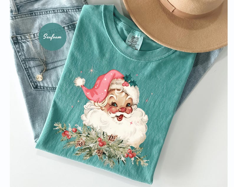 May include: Seafoam green t-shirt featuring a vintage Santa Claus graphic with a pink hat and white beard. The Santa holds a sprig of pine with red berries. The shirt is paired with blue jeans and a tan hat.