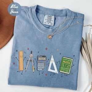 May include: A blue jean coloured t-shirt featuring a graphic print of five anthropomorphic school supplies including a ruler, compass, calculator, triangle, and a green book labelled MATH, all surrounded by small red and blue star illustrations.