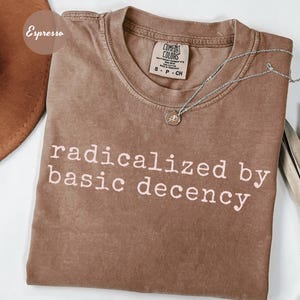 May include: A brown t-shirt with the text "radicalized by basic decency" printed in white. The shirt has a distressed look and is made of a soft, comfortable fabric.