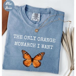 Pode incluir: Uma camiseta azul claro Comfort Colors com o texto "THE ONLY ORANGE MONARCH I WANT" e um gr&aacute;fico de borboleta monarca. Um colar de ouro est&aacute; sobre a camiseta. A camiseta tem gola redonda e mangas curtas.