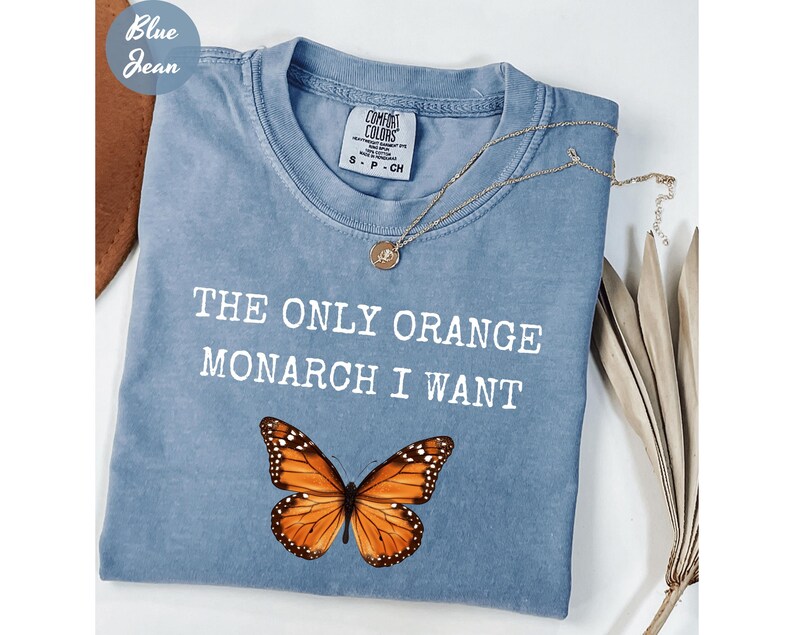 Pode incluir: Uma camiseta azul claro Comfort Colors com o texto "THE ONLY ORANGE MONARCH I WANT" e um gr&aacute;fico de borboleta monarca. Um colar de ouro est&aacute; sobre a camiseta. A camiseta tem gola redonda e mangas curtas.