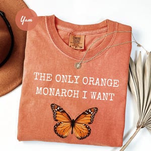 May include: Coral-colored t-shirt with the text "THE ONLY ORANGE MONARCH I WANT" and a monarch butterfly graphic. A gold necklace and a brown hat are also visible.