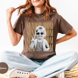 May include: A brown short-sleeve t-shirt featuring a black and white photograph of a child wearing heart-shaped sunglasses and making a peace sign. The text SENIOR MOM is repeated in gold behind the photo, with the name Amelia written below.
