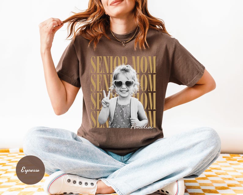 May include: A brown short-sleeve t-shirt featuring a black and white photograph of a child wearing heart-shaped sunglasses and making a peace sign. The text SENIOR MOM is repeated in gold behind the photo, with the name Amelia written below.