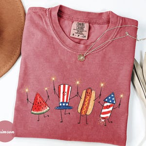 K&ouml;nnte beinhalten: Ein rotes Comfort Colors T-Shirt mit einer skurrilen Illustration einer Wassermelonenscheibe, einem patriotischen Hut, einem Hotdog und einem Feuerwerk, die jeweils einen Wunderkerzenhalter halten. Eine goldene Halskette und ein brauner Hut sind ebenfalls sichtbar.
