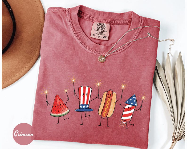 K&ouml;nnte beinhalten: Ein rotes Comfort Colors T-Shirt mit einer skurrilen Illustration einer Wassermelonenscheibe, einem patriotischen Hut, einem Hotdog und einem Feuerwerk, die jeweils einen Wunderkerzenhalter halten. Eine goldene Halskette und ein brauner Hut sind ebenfalls sichtbar.