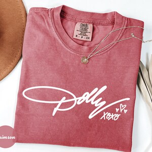 May include: A crimson short-sleeved t-shirt featuring the white script text Dolly XOXO with two small hearts. The shirt has a crew neckline and a Comfort Colors brand label on the inside collar. A gold-toned necklace with a small circular pendant rests on the fabric.