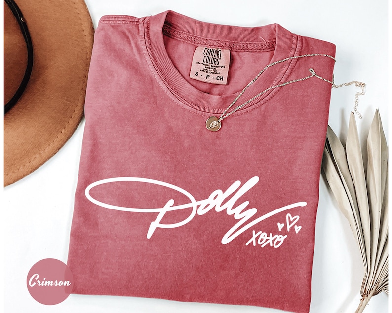 May include: A crimson short-sleeved t-shirt featuring the white script text Dolly XOXO with two small hearts. The shirt has a crew neckline and a Comfort Colors brand label on the inside collar. A gold-toned necklace with a small circular pendant rests on the fabric.