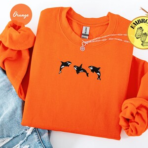 May include: Orange sweatshirt with three embroidered orca whales in black and white. The sweatshirt has a crew neck and long sleeves. A silver necklace with a sun pendant is visible. The word "Embroidery" is in a yellow circle.