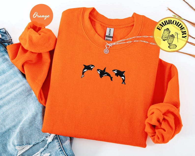 May include: Orange sweatshirt with three embroidered orca whales in black and white. The sweatshirt has a crew neck and long sleeves. A silver necklace with a sun pendant is visible. The word "Embroidery" is in a yellow circle.