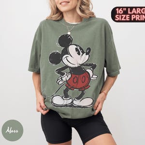 Puede incluir: Camiseta verde oliva con un gran gr&aacute;fico de Mickey Mouse. El personaje de dibujos animados est&aacute; en negro, blanco y rojo. El texto "16" LARGE SIZE PRINT" est&aacute; en un &oacute;valo negro en la esquina superior derecha.