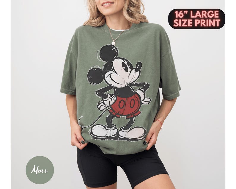 Puede incluir: Camiseta verde oliva con un gran gr&aacute;fico de Mickey Mouse. El personaje de dibujos animados est&aacute; en negro, blanco y rojo. El texto "16" LARGE SIZE PRINT" est&aacute; en un &oacute;valo negro en la esquina superior derecha.