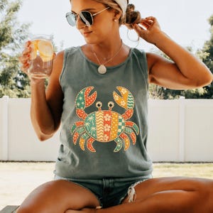 May include: A blue-green tank top featuring a colorful crab design. The crab is decorated with various patterns in shades of red, green, yellow, and orange. The tank top is sleeveless and appears to be made of a soft material.