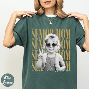 May include: A dark green short-sleeve t-shirt featuring a black and white photograph of a child wearing heart-shaped sunglasses and making a peace sign. The text SENIOR MOM is repeated in gold behind the photo, with the name Amelia in script.