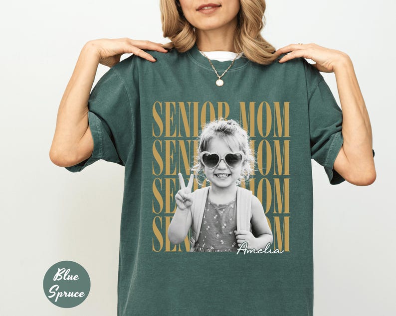 May include: A dark green short-sleeve t-shirt featuring a black and white photograph of a child wearing heart-shaped sunglasses and making a peace sign. The text SENIOR MOM is repeated in gold behind the photo, with the name Amelia in script.