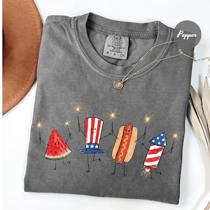 K&ouml;nnte beinhalten: Graues Comfort Colors T-Shirt mit einer Grafik einer Wassermelonenscheibe, einem Hotdog und einem Feuerwerk, das Wunderkerzen h&auml;lt. Das Shirt hat auch einen rot-wei&szlig;-blauen Uncle Sam Hut. Eine Halskette mit Anh&auml;nger befindet sich auf dem Shirt.