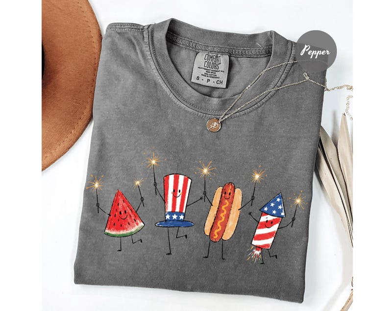 K&ouml;nnte beinhalten: Graues Comfort Colors T-Shirt mit einer Grafik einer Wassermelonenscheibe, einem Hotdog und einem Feuerwerk, das Wunderkerzen h&auml;lt. Das Shirt hat auch einen rot-wei&szlig;-blauen Uncle Sam Hut. Eine Halskette mit Anh&auml;nger befindet sich auf dem Shirt.