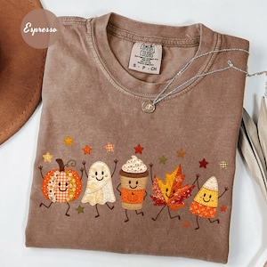May include: An espresso-coloured short-sleeve t-shirt featuring a graphic print of a smiling pumpkin, ghost, coffee cup, maple leaf, and candy corn. The design includes small colourful stars and the text Espresso is visible in the upper left corner.