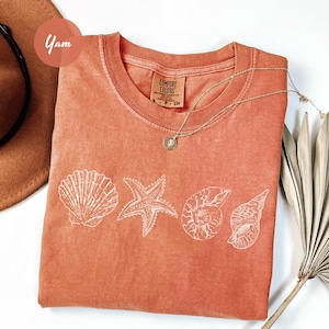 May include: Coral-colored Comfort Colors t-shirt featuring a white seashell design. The design includes a scallop shell, starfish, and two different spiral shells. A silver necklace and a brown hat are also in the image.