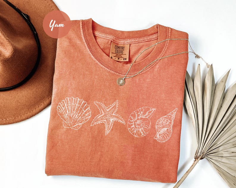 May include: Coral-colored Comfort Colors t-shirt featuring a white seashell design. The design includes a scallop shell, starfish, and two different spiral shells. A silver necklace and a brown hat are also in the image.