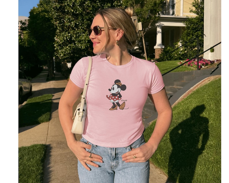 Pu&ograve; includere: Una maglietta rosa chiaro con una grafica classica di Minnie Mouse. Il personaggio dei cartoni animati indossa un vestito a pois e un fiocco. La maglietta &egrave; a maniche corte e abbinata a jeans blu. Una borsa bianca &egrave; indossata a tracolla.