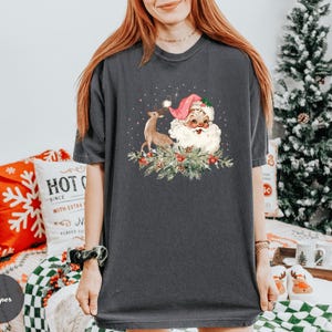 May include: Dark gray t-shirt featuring a vintage Santa Claus illustration with a reindeer, berries, and greenery. The Santa Claus is wearing a pink hat and has a white beard. The shirt is a comfortable, casual style, perfect for the holiday season.