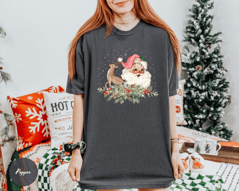 May include: Dark gray t-shirt featuring a vintage Santa Claus illustration with a reindeer, berries, and greenery. The Santa Claus is wearing a pink hat and has a white beard. The shirt is a comfortable, casual style, perfect for the holiday season.