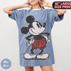 Puede incluir: Una camiseta vaquera azul con un gran gr&aacute;fico de Mickey Mouse. El personaje de dibujos animados es en negro, blanco y rojo. La camiseta tiene una impresi&oacute;n de gran tama&ntilde;o de 40,6 cm.
