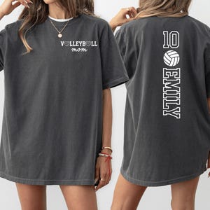 May include: Dark gray t-shirt with the text "VOLLEYBALL mom" on the front and "10 EMILY" with a volleyball graphic on the back. The shirt has a crew neck and short sleeves. The shirt is a comfortable, casual style.