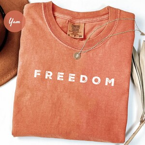 May include: A coral-colored t-shirt with the word "FREEDOM" printed in white block letters. A gold necklace with a pendant rests on the shirt. A brown hat and dried palm leaves are in the background. The shirt is made by Comfort Colors.
