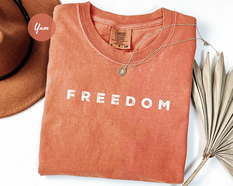 May include: A coral-colored t-shirt with the word "FREEDOM" printed in white block letters. A gold necklace with a pendant rests on the shirt. A brown hat and dried palm leaves are in the background. The shirt is made by Comfort Colors.
