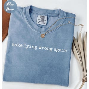 May include: A blue jean colored t-shirt with the phrase "make lying wrong again" in white text. The shirt has a crew neck and a small tag on the upper back. A gold necklace with a pendant is draped across the shirt.