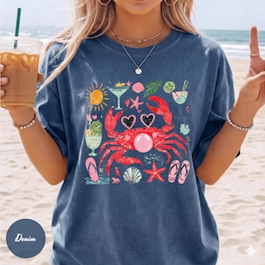 May include: Denim blue t-shirt featuring a colorful beach-themed graphic. The design includes a red crab wearing heart-shaped sunglasses, cocktails, seashells, and a sun. The shirt is a relaxed fit.