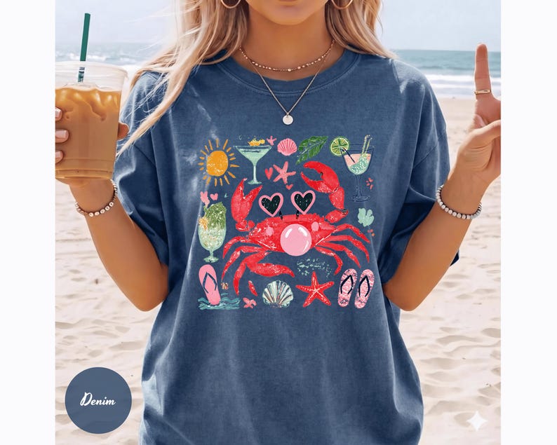May include: Denim blue t-shirt featuring a colorful beach-themed graphic. The design includes a red crab wearing heart-shaped sunglasses, cocktails, seashells, and a sun. The shirt is a relaxed fit.