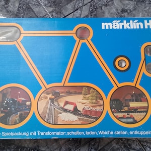 Peut inclure: Une boîte de train miniature Märklin HO bleue avec des lignes graphiques orange reliant trois fenêtres circulaires montrant des locomotives à vapeur et des wagons sur des rails. Le texte indique Märklin HO et große Spielpackung mit Transformator; schalten, laden, Weiche stellen, entkuppeln, rangieren.
