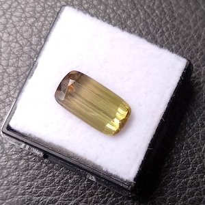 May include: A rectangular, faceted gemstone with a gradient color transition from deep brownish-gold at one end to a lighter, translucent yellow at the other. The stone is displayed resting on a white foam insert inside a small black plastic case.