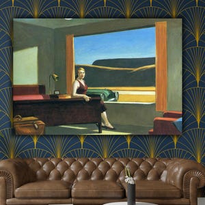 Puede incluir: Una impresión enmarcada de "Hotel Room" de Edward Hopper cuelga sobre un sofá de cuero marrón. La pintura representa a una mujer con un vestido granate en una habitación de hotel, con una maleta y un coche visibles a través de la ventana. La habitación tiene un escritorio y una lámpara.